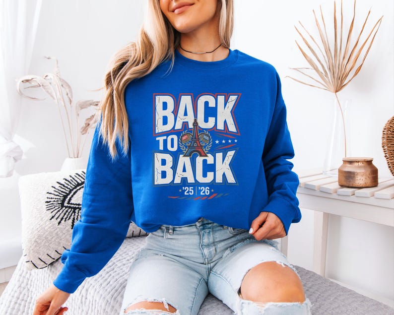 A woman wearing a blue sweatshirt with the text "BACK TO BACK" is sitting on a bed in a room with various decorative items.