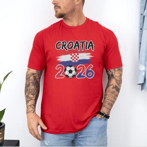 A man wearing a red t-shirt with the text "Croatia 2026" and a soccer ball graphic stands in front of a white wall.