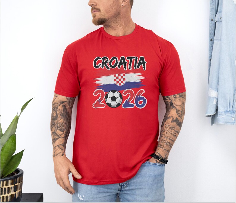 A man wearing a red t-shirt with the text "Croatia 2026" and a soccer ball graphic stands in front of a white wall.