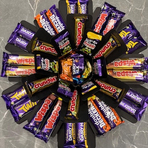 May include: A black box shaped like a flower with different chocolate bars in each section. The chocolate bars include Dairy Milk, Wispa, Twirl, Crunchie, Freddo, and Double Decker.