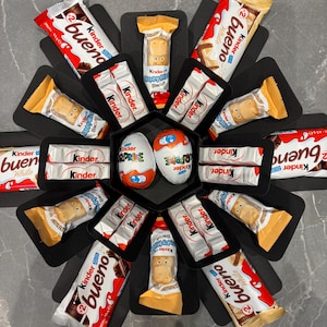 May include: A black explosion box filled with various Kinder chocolate products, including Kinder Bueno, Kinder Surprise, and Kinder Happy Hippo biscuits.