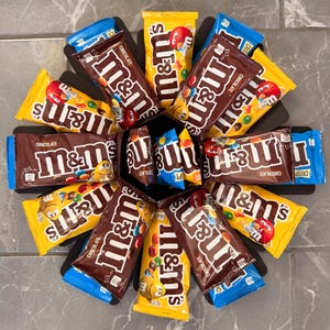 May include: A top-down view of a black box surrounded by M&M's chocolate bars and bags. The bars are brown with the M&M's logo, and the bags are yellow and blue. The candy is arranged in a radial pattern.