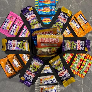 May include: A black box filled with a variety of chocolate bars and sweets. The box is arranged in a circular pattern with the sweets facing outwards. The sweets include brands like Freddo, Maoam, Haribo, and Drumstick.