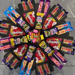 May include: A black gift box, arranged in a circular pattern, filled with a variety of wrapped sweets and chocolate bars. Brands include Skittles, Freddo, Wispa, Vimto, and Refresher. Each compartment contains different treats.
