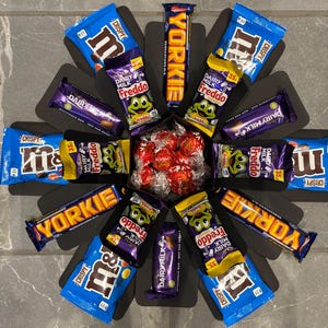 May include: A black circular box with a flower design, filled with various chocolate bars, including Yorkie, Dairy Milk, Freddo, and M&Ms. The box also contains a small bag of red chocolate sweets.