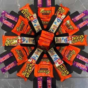 May include: A black gift box arrangement filled with assorted sweets. The box contains Reese's Peanut Butter Cups, Reese's Sticks, Milky Way bars, and Vimto candies. The sweets are arranged in a starburst pattern.