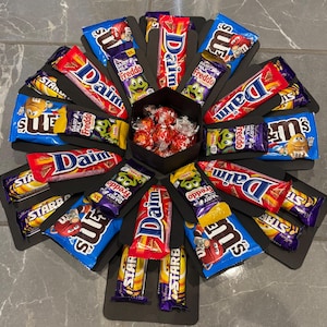 May include: A black gift box arrangement filled with a variety of sweets. The box features Dairy Milk Freddo, Daim, Starburst, and M&M's chocolate bars. A central section contains wrapped chocolate balls.