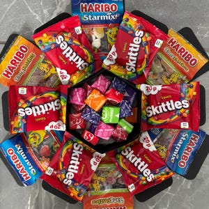 May include: A hexagonal box filled with various bags of sweets. The box is black and the bags are red, yellow, and blue. The sweet bags are labelled with the brand names "HARIBO" and "Skittles".