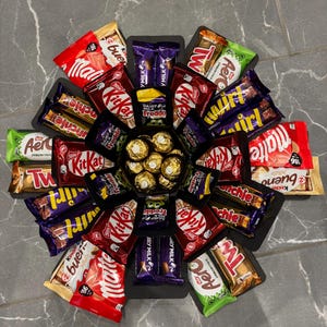 May include: A black box filled with various chocolate bars, including Twix, KitKat, Aero, and Milky Way. The box is arranged in a circular pattern with the chocolate bars facing outwards. There are also some round chocolate candies in the centre of the box.