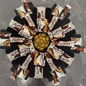 May include: A black gift box filled with chocolate bars. The box is shaped like a flower with 12 chocolate bars arranged around a centre filled with chocolate eggs. The chocolate bars are Galaxy, Flutes, and Galaxy Minstrels. The box is on a grey surface.