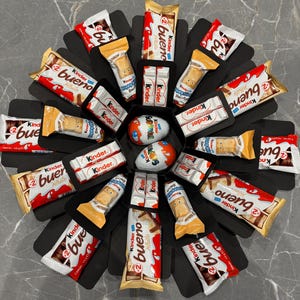 May include: A circular arrangement of Kinder chocolate products, including Bueno bars, Happy Hippo biscuits, and Surprise eggs, in a black box. The packaging features red, white, and gold colours, typical of the Kinder brand.