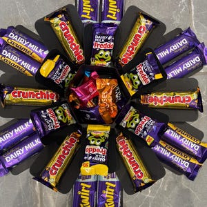 May include: A black hexagonal box filled with various chocolate bars, including Cadbury Dairy Milk Caramel, Twirl, Crunchie, and Dairy Milk Freddo. The box is surrounded by a black border.