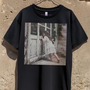May include: Black t-shirt featuring a square graphic of a young girl in a white dress peering through a weathered door. The text "violent femmes" is above the image. The shirt is hanging on a purple hanger.