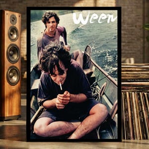 May include: A framed poster featuring the band Ween, showing two individuals in a small boat on water. One person sits in the foreground lighting a cigarette, while the other sits behind them. The word Ween is printed in white script at the top.