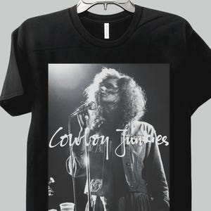 May include: Black t-shirt featuring a black and white image of a person singing into a microphone. The image has the words "Cowboy Junkies" in white script. The shirt is hanging on a metal hanger.