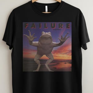 May include: Black t-shirt featuring a graphic of a frog with its arms raised against a sunset backdrop. The word "FAILURE" is printed in red block letters above the frog, with "magnified" in smaller text below.