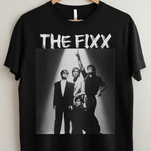 May include: Black t-shirt featuring the band name "THE FIXX" in white, above a black and white image of the band members. The band members are in various poses, with one pointing upwards.