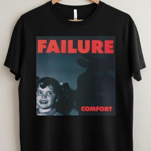 May include: Black t-shirt featuring a graphic design with the word "FAILURE" in red block letters above a vintage image of a young girl and a silhouette. The word "COMFORT" is below the image.
