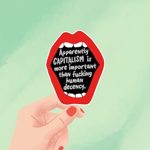 May include: A sticker with a red mouth graphic, holding the text "Apparently CAPITALISM is more important than fucking human decency." The sticker is held by a hand with red nail polish against a light green background.
