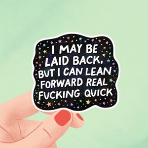 Puede incluir: Una pegatina negra con texto blanco que dice: "I MAY BE LAID BACK, BUT I CAN LEAN FORWARD REAL FUCKING QUICK". La pegatina tiene un borde blanco y está decorada con estrellas de colores.