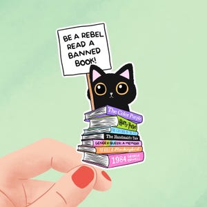 Könnte beinhalten: Ein Aufkleber mit einer schwarzen Katze, die ein Schild mit der Aufschrift "BE A REBEL READ A BANNED BOOK!" hält. Die Katze sitzt auf einem Stapel Bücher, darunter "1984" und "Harry Potter".