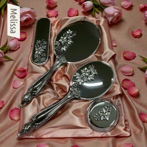 以下が含まれることがあります： 手鏡、小さな丸いコンパクト、ブラシを含む、花の装飾が施された銀色の化粧台セット。 セットはピンクのサテン生地の上に展示されており、背景にはピンクのバラの花びらとバラがあります。 「Melissa」という言葉が見えます。