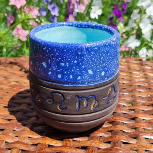 May include: A handcrafted ceramic cup with a blue interior and a speckled blue exterior. The lower portion is brown with zodiac symbols. The cup sits on a woven surface, with a backdrop of colorful flowers.