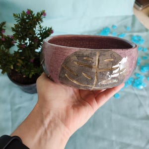 May include: A handmade ceramic bowl with a crackled, pink exterior and a brown interior. The bowl features a leaf design in shades of gray and brown. The bowl is held in a hand, with a small plant in the background.