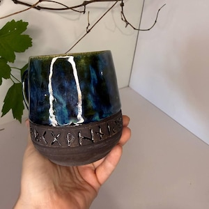May include: A ceramic mug featuring a glossy, deep blue and green marbled glaze on the upper portion and a matte, dark brown textured base. The lower section is embossed with a horizontal band of carved runic symbols encircling the vessel.