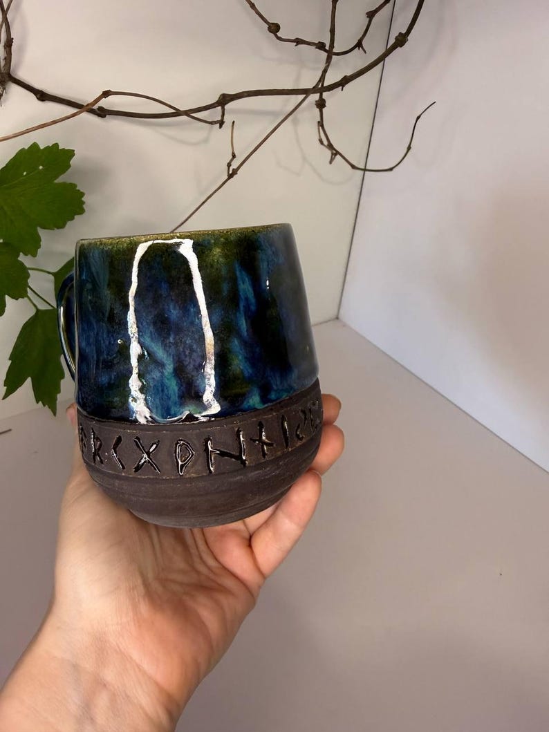 May include: A ceramic mug featuring a glossy, deep blue and green marbled glaze on the upper portion and a matte, dark brown textured base. The lower section is embossed with a horizontal band of carved runic symbols encircling the vessel.