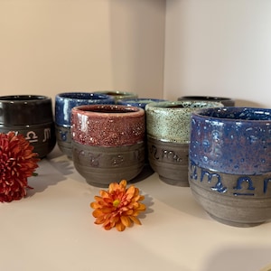 Op de afbeelding: Een verzameling handgemaakte keramische bekers in verschillende kleuren, waaronder blauw, rood en groen, met een donkerbruine basis. Elke beker heeft een dierenriemteken en is ongeveer 10 cm hoog. Twee bloemen in rood- en oranje tinten staan op de voorgrond.