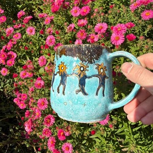 May include: A light blue ceramic mug featuring a hand-painted design of stylised figures with sun-like heads holding hands in a circle. The mug has a textured, mottled glaze finish with a dark brown rim and small star and circle symbols near the base.