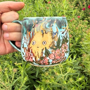 May include: A ceramic mug with a light blue glaze featuring a hand-painted yellow flame character with large eyes and a small mouth, surrounded by pink flowers and green leaves. The top rim has a dark teal drip effect, and a curved handle is attached.