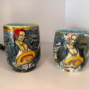Puede incluir: Dos vasos de cerámica con una ilustración de dibujos animados de un personaje con cabello rojo, sosteniendo un sombrero amarillo. Los vasos tienen un interior azul y están decorados con un fondo detallado. Los vasos están sobre una superficie blanca.