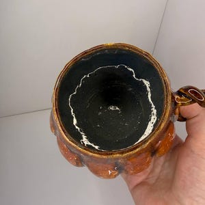 May include: A ceramic mug shaped like a pumpkin with a glossy orange exterior and a dark black interior. The rim features a distinct white drip pattern along the inner edge. The mug includes a twisted handle on the side, showcasing a textured, hand-crafted appearance with a rounded, segmented body design.