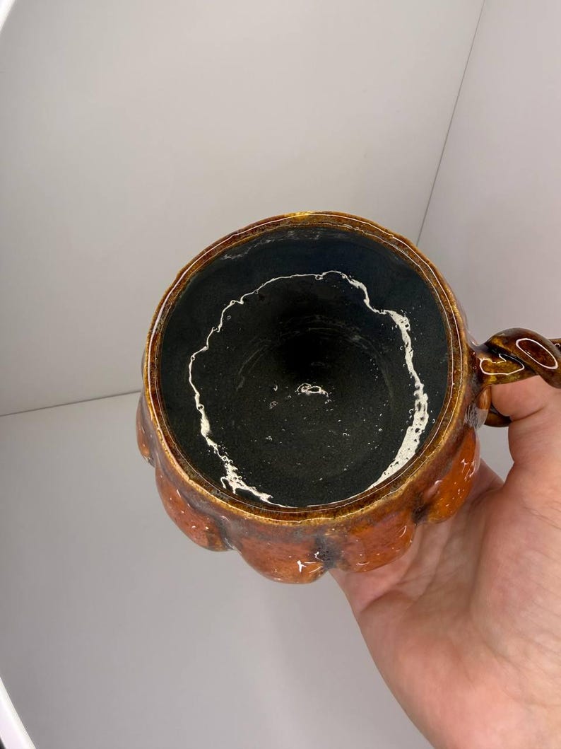 May include: A ceramic mug shaped like a pumpkin with a glossy orange exterior and a dark black interior. The rim features a distinct white drip pattern along the inner edge. The mug includes a twisted handle on the side, showcasing a textured, hand-crafted appearance with a rounded, segmented body design.