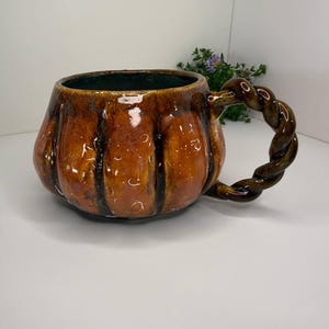 May include: A glossy, pumpkin-shaped ceramic mug with a warm, mottled orange and brown glaze. The mug features vertical ridges along its rounded body and a distinctive, thick handle crafted in a twisted, rope-like design with a dark brown finish.