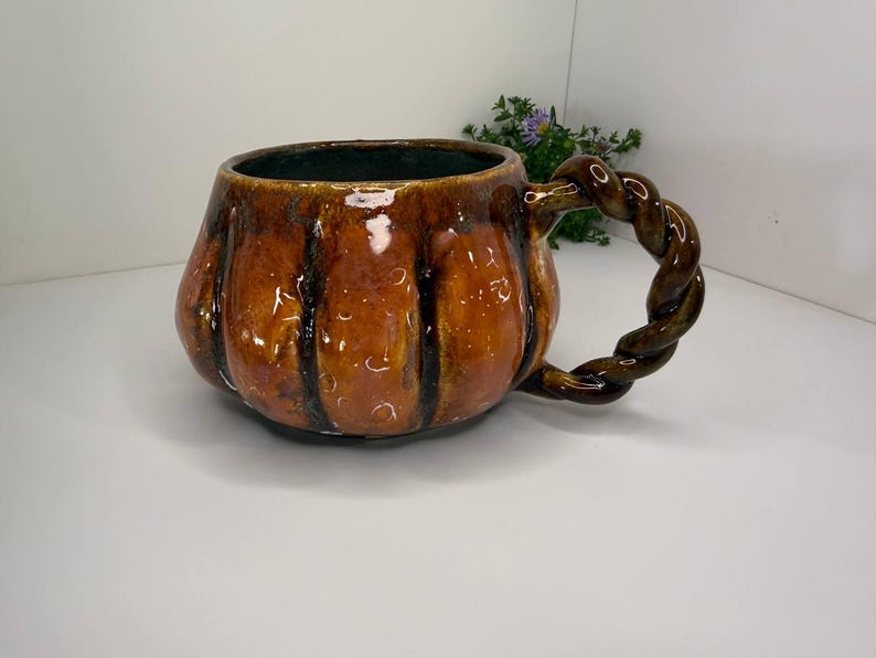 May include: A glossy, pumpkin-shaped ceramic mug with a warm, mottled orange and brown glaze. The mug features vertical ridges along its rounded body and a distinctive, thick handle crafted in a twisted, rope-like design with a dark brown finish.