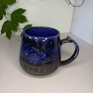 May include: A ceramic mug with a deep blue and black mottled glaze on the upper portion and a dark, unglazed base featuring a band of embossed runic characters. The interior is a solid, vibrant blue, and the handle is curved and dark in color.