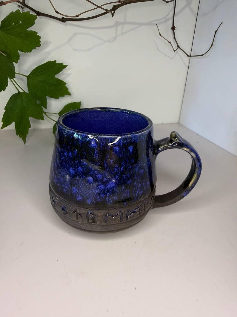 May include: A ceramic mug with a deep blue and black mottled glaze on the upper portion and a dark, unglazed base featuring a band of embossed runic characters. The interior is a solid, vibrant blue, and the handle is curved and dark in color.