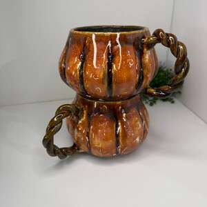 May include: Two stacked ceramic mugs shaped like pumpkins with a glossy orange and brown glaze. Each mug features vertical ridges and a dark brown twisted rope-style handle. The mugs have a smooth, reflective finish and a rounded, bulbous body.