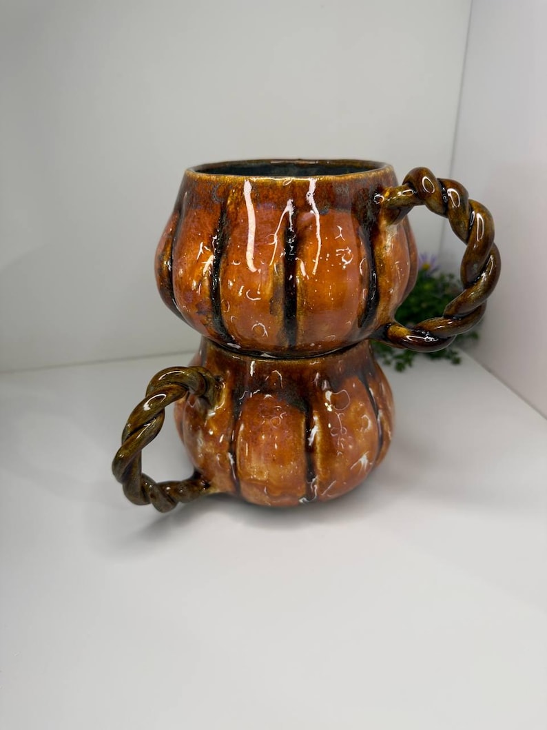 May include: Two stacked ceramic mugs shaped like pumpkins with a glossy orange and brown glaze. Each mug features vertical ridges and a dark brown twisted rope-style handle. The mugs have a smooth, reflective finish and a rounded, bulbous body.