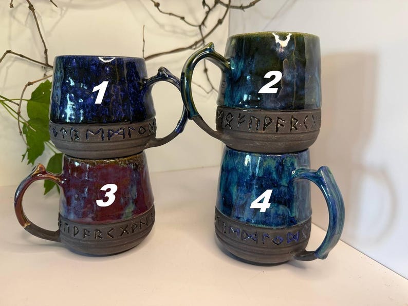 May include: Four ceramic mugs with handles, each featuring a dark, unglazed base band embossed with runic characters. Mug 1 is deep blue, mug 2 is dark green and blue, mug 3 is a mix of red and purple, and mug 4 is a vibrant blue and teal blend.