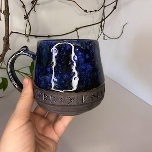 May include: A ceramic mug with a glossy, mottled dark blue and black upper section featuring a white outlined shape. The lower portion is matte dark gray with a band of carved runic characters. It has a curved handle and a rounded base.