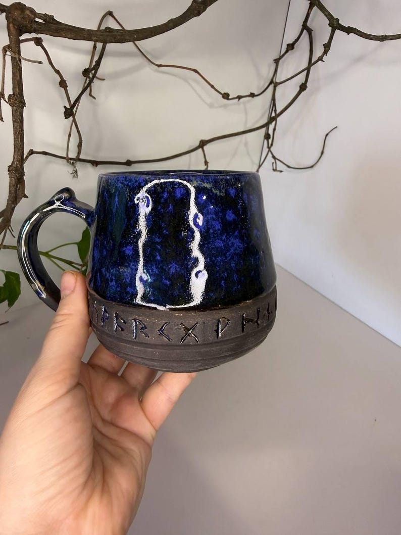 May include: A ceramic mug with a glossy, mottled dark blue and black upper section featuring a white outlined shape. The lower portion is matte dark gray with a band of carved runic characters. It has a curved handle and a rounded base.