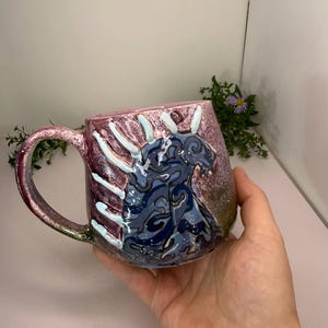 Op de afbeelding: Een keramische mok met een glanzend roze en paars gespikkeld glazuur, voorzien van een donkerblauw abstract wervelpatroon aan de zijkant. De mok heeft een gebogen handvat en een reeks verhoogde, lichtblauwe verticale ribbels langs de bovenrand.
