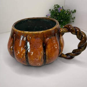 May include: A ceramic mug with a glossy, pumpkin-orange glaze and vertical ridges. The mug features a dark interior and a distinctive, thick, twisted rope-style handle in a matching dark brown and orange finish, creating a textured appearance.