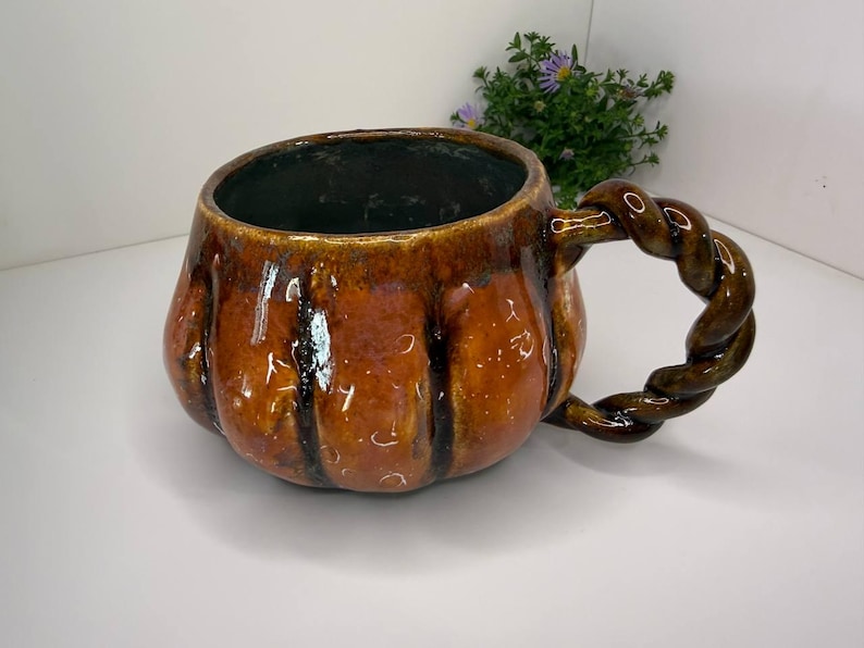 May include: A ceramic mug with a glossy, pumpkin-orange glaze and vertical ridges. The mug features a dark interior and a distinctive, thick, twisted rope-style handle in a matching dark brown and orange finish, creating a textured appearance.