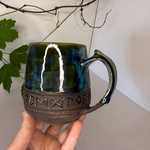 May include: A ceramic mug featuring a glossy, deep blue and green glazed upper section and a dark, textured matte brown base. The lower portion is embossed with a series of runic characters encircling the body. It has a curved handle.