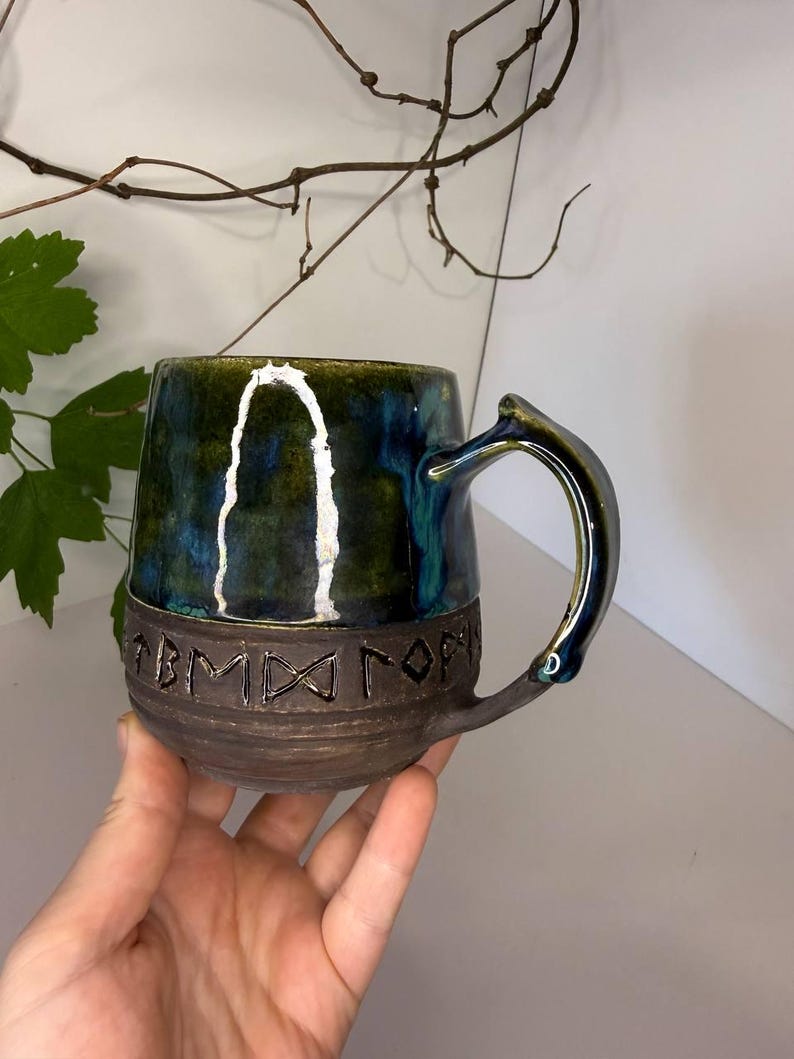 May include: A ceramic mug featuring a glossy, deep blue and green glazed upper section and a dark, textured matte brown base. The lower portion is embossed with a series of runic characters encircling the body. It has a curved handle.
